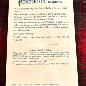 Pu&ograve; includere: Un foglio di carta bianco con il logo e il testo di Pendleton Blankets, su una coperta a fantasia rossa e nera. Il testo descrive la qualit&agrave; delle coperte, le istruzioni per la cura e la storia dell'azienda.