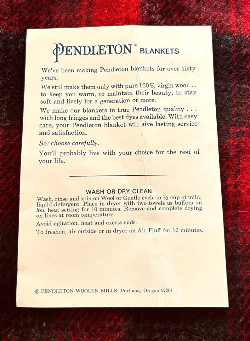 Pu&ograve; includere: Un foglio di carta bianco con il logo e il testo di Pendleton Blankets, su una coperta a fantasia rossa e nera. Il testo descrive la qualit&agrave; delle coperte, le istruzioni per la cura e la storia dell'azienda.
