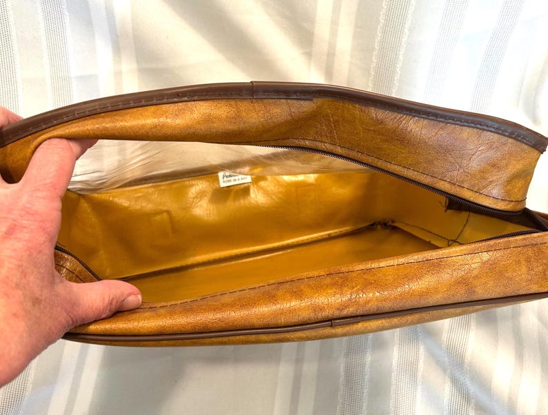 Pu&ograve; includere: Vista aperta di una valigia marrone vintage con interno dorato. La valigia ha un esterno strutturato con finiture marrone scuro e chiusura con cerniera. L'interno &egrave; di colore oro lucido. La valigia &egrave; aperta, rivelando l'interno.
