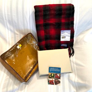 Pu&ograve; includere: Una sciarpa a quadri rossi e neri con bordo a frange, un cuscino color crema e una borsa marrone con cerniera. &Egrave; presente anche una piccola carta con immagini di prodotti simili. La sciarpa ha un'etichetta con una scena di montagna.