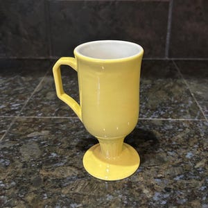 May include: A yellow ceramic mug with a white interior. The mug has a pedestal base and a square handle. The mug is a solid yellow color and is sitting on a dark countertop.