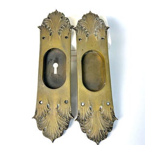 May include: Two antique brass door handle plates. Each plate has an oval opening, a keyhole, and ornate leaf-like designs. The plates are rectangular with rounded tops and bottoms, and are set against a white background.