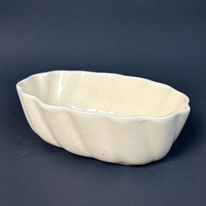 May include: Cream-colored, oval-shaped ceramic dish with a wavy, textured exterior. The dish has a crackled surface and is likely vintage. The design is simple and elegant, suitable for serving or display.