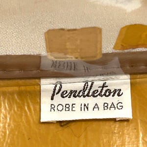 Pu&ograve; includere: Primo piano di un'etichetta con la scritta "Pendleton ROBE IN A BAG" in nero su sfondo bianco. L'etichetta &egrave; attaccata a un tessuto color senape. Lo sfondo &egrave; beige chiaro.