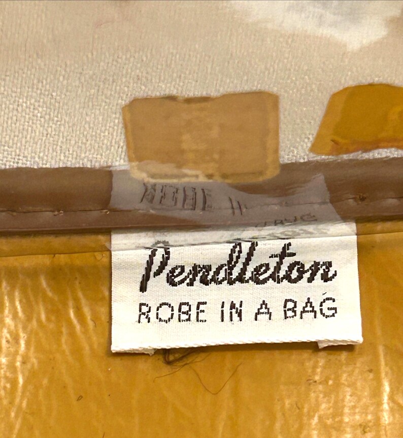 Pu&ograve; includere: Primo piano di un'etichetta con la scritta "Pendleton ROBE IN A BAG" in nero su sfondo bianco. L'etichetta &egrave; attaccata a un tessuto color senape. Lo sfondo &egrave; beige chiaro.