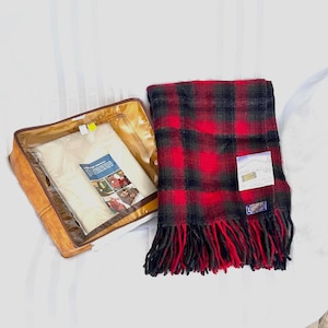 Pu&ograve; includere: Una coperta di lana a quadri rossi e neri con bordo a frange, accanto a una custodia in similpelle color cuoio con una finestra di plastica trasparente. La coperta ha un'etichetta attaccata. La custodia contiene una coperta bianca.