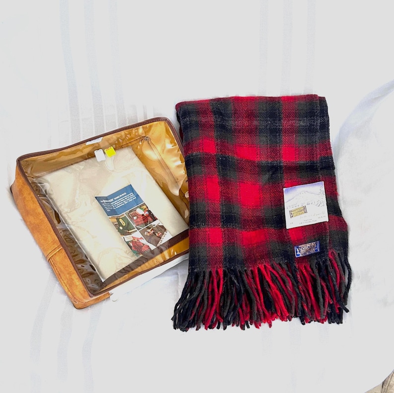 Pu&ograve; includere: Una coperta di lana a quadri rossi e neri con bordo a frange, accanto a una custodia in similpelle color cuoio con una finestra di plastica trasparente. La coperta ha un'etichetta attaccata. La custodia contiene una coperta bianca.