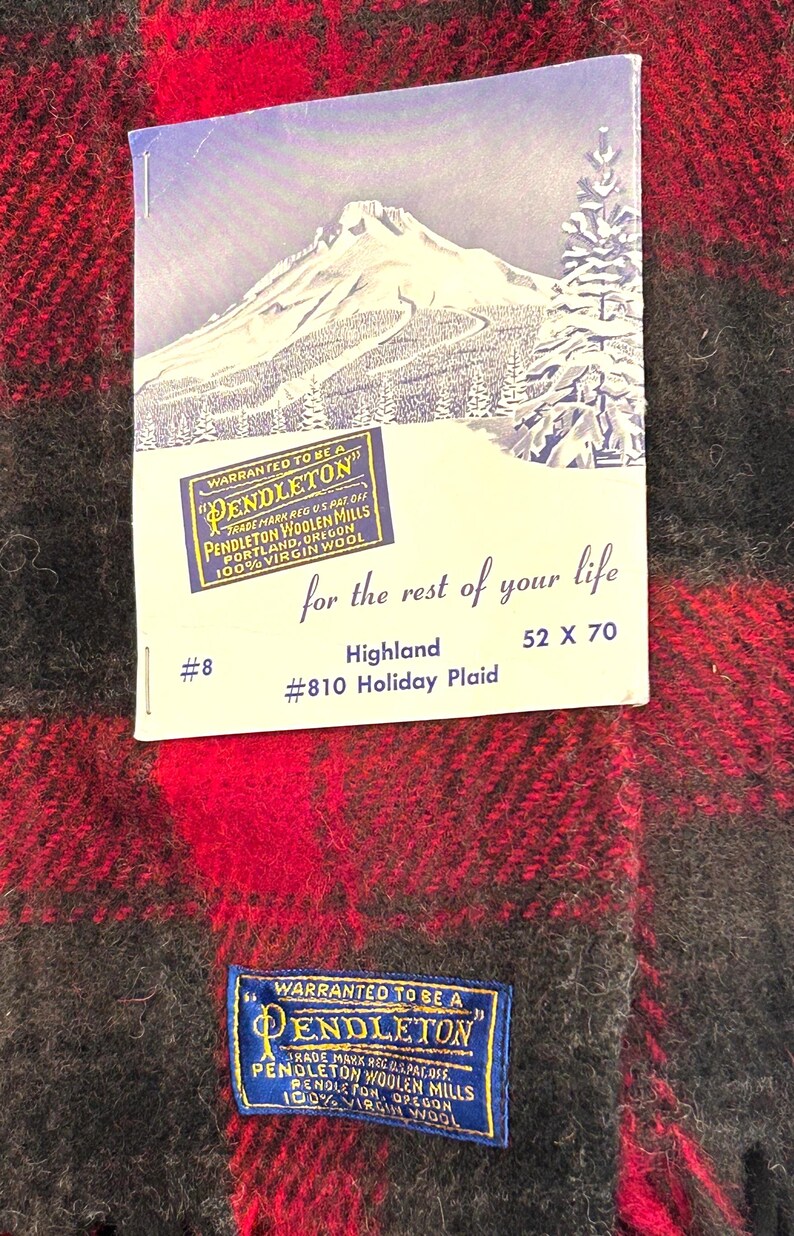 Pu&ograve; includere: Una coperta a quadri rossi e neri con un'etichetta che recita "Pendleton Woolen Mills". La coperta misura 132 x 178 cm e presenta il testo "Highland #810 Holiday Plaid". L'etichetta ha anche un'illustrazione di una montagna e alberi.