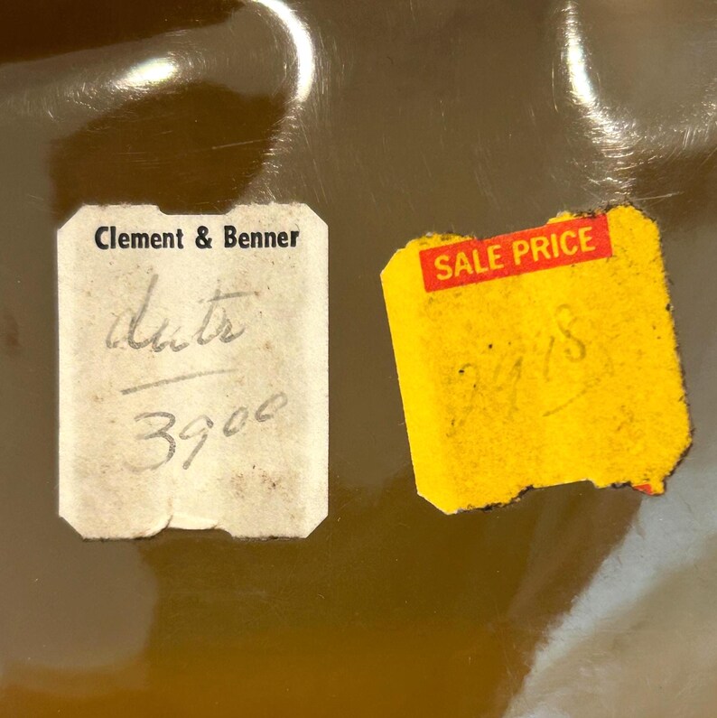 Pu&ograve; includere: Primo piano di due vecchie etichette di prezzo su una superficie riflettente color bronzo. Un'etichetta &egrave; bianca con il testo "Clement & Benner" e testo e numeri scritti a mano. L'altra &egrave; gialla con "SALE PRICE" in rosso e numeri scritti a mano.