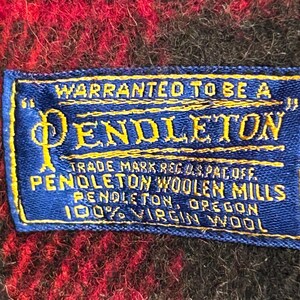 Pu&ograve; includere: Primo piano di un'etichetta Pendleton blu e gialla su un tessuto a quadri rosso e nero. L'etichetta recita "WARRANTED TO BE A PENDLETON" e include dettagli sul marchio, il marchio e il materiale: 100% lana vergine.