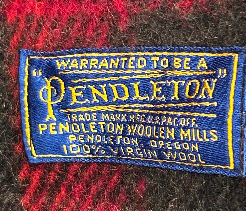 Pu&ograve; includere: Primo piano di un'etichetta Pendleton blu e gialla su un tessuto a quadri rosso e nero. L'etichetta recita "WARRANTED TO BE A PENDLETON" e include dettagli sul marchio, il marchio e il materiale: 100% lana vergine.