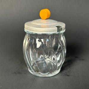 May include: A clear glass jar with a silver lid and an orange knob. The jar has a swirled design and is empty. The lid has a small opening. The jar is sitting on a black surface.