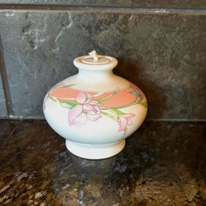 May include: A white ceramic oil lamp with a floral design in pink, peach, and green. The lamp has a round body, a small base, and a lid with a wick. The lamp is likely used for aromatherapy or decorative purposes.