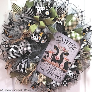 May include: A decorative wreath featuring black, white, and olive green mesh ribbons with cow print, gingham, and daisy patterns. A rectangular sign in the center displays three cows wearing floral crowns and the text LIVE LIKE SOMEONE LEFT THE GATE OPEN.