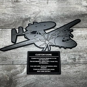 May include: A metal cutout of a C-2 Greyhound aircraft, a military transport plane. The cutout is silver and has a black background. The cutout is mounted on a wooden surface. The cutout comes with a custom name and message.