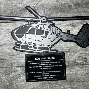May include: A black metal silhouette of a helicopter with a silver finish. The helicopter is mounted on a black leather plaque with the text "CUSTOM NAME" and a message about adding a custom message, logo, rank, or insignia.
