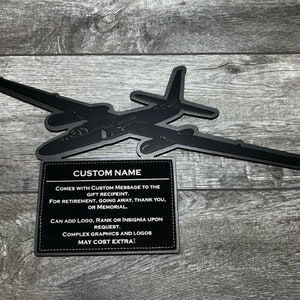 May include: Black silhouette of a U-2 spy plane with a black and white sign that reads "CUSTOM NAME" and includes the text "COMES WITH CUSTOM MESSAGE TO THE GIFT RECIPIENT. FOR RETIREMENT, GOING AWAY, THANK YOU, OR MEMORIAL. CAN ADD LOGO, RANK OR INSIGNIA UPON REQUEST. COMPLEX GRAPHICS AND LOGOS MAY COST EXTRA!"