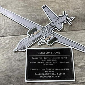 May include: A gray and white airplane sticker with a black rectangle below it. The rectangle has the words "CUSTOM NAME" at the top and text below it. The airplane has a propeller and is carrying missiles.