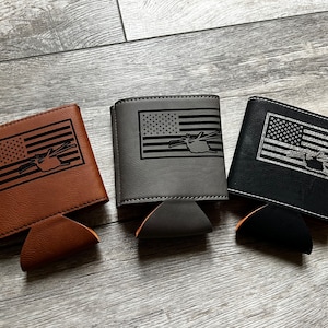 May include: Three different colored can coolers with an American flag and a helicopter design. The coolers are brown, gray, and black.