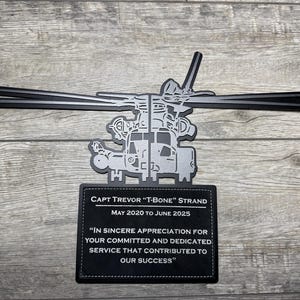 May include: A black and silver metal helicopter-shaped plaque with a rectangular nameplate. The nameplate reads "CAPT TREVOR 'T-BONE' STRAND MAY 2020 TO JUNE 2025" and an appreciation message. The plaque is on a wooden surface.