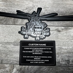 May include: Black metal silhouette of a helicopter with a black leather plaque underneath. The plaque has white text that reads "CUSTOM NAME", "COMES WITH CUSTOM MESSAGE TO THE GIFT RECIPIENT", "FOR RETIREMENT, GOING AWAY, THANK YOU, OR MEMORIAL", "CAN ADD LOGO, RANK OR INSIGNIA UPON REQUEST", "COMPLEX GRAPHICS AND LOGOS MAY COST EXTRA!"