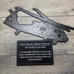 May include: A dark gray metal helicopter silhouette with a matching plaque. The plaque reads "CAPT TREVOR 'T-BONE' STRAND MAY 2020 TO JUNE 2025" and a dedication message. The helicopter has detailed features.