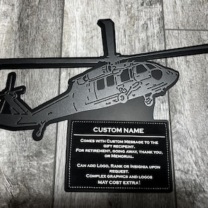 May include: Black metal silhouette of a helicopter with a custom name plate. The plate has text that reads "CUSTOM NAME", "COMES WITH CUSTOM MESSAGE TO THE GIFT RECIPIENT. FOR RETIREMENT, GOING AWAY, THANK YOU, OR MEMORIAL. CAN ADD LOGO, RANK OR INSIGNIA UPON REQUEST. COMPLEX GRAPHICS AND LOGOS MAY COST EXTRA!"