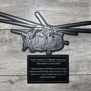 May include: A silver metal plaque with a black leather background. The plaque features a silhouette of a helicopter and text that reads "CAPT TREVOR "T-BONE" STRAND MAY 2020 TO JUNE 2025 "IN SINCERE APPRECIATION FOR YOUR COMMITTED AND DEDICATED SERVICE THAT CONTRIBUTED TO OUR SUCCESS"