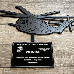 May include: A black metal plaque with a silver helicopter silhouette and the text "Maj Scott "Tool" Thomson VMM-169 Thanks for your dedication and hard work. Jan 2020 to May 2022 "Semper Fi""