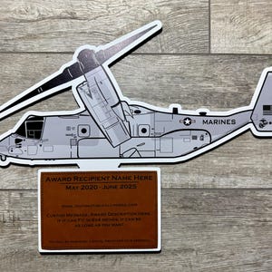 May include: A white and gray model of a V-22 Osprey aircraft with the words "MARINES" and "00" printed on the side. The model is mounted on a brown plaque with the text "AWARD RECIPIENT NAME HERE MAY 2020 - JUNE 2025" and a custom message area.
