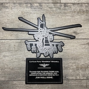 May include: A black and silver metal wall art piece featuring a detailed helicopter silhouette above a rectangular plaque. The plaque displays the text: Captain Pete "Maverick" Mitchell, an emblem, and the message: Thanks for training those kids, completing the mission, and bringing everyone home safely. Job well done.