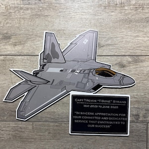 May include: A gray F-22 Raptor fighter jet sticker with a black and white border. A black rectangle with white text reads "Capt Trevor 'T-Bone' Strand" and a message of appreciation for service.