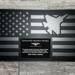 May include: A black and silver American flag with a fighter jet silhouette and a plaque that reads "Captain Pete "Maverick" Mitchell" and "Thanks for training those kids, completing the mission, and bringing everyone home safely. Job well done."