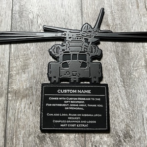 May include: Black metal silhouette of a helicopter with a custom name plate. The plate has text that reads "CUSTOM NAME", "COMES WITH CUSTOM MESSAGE TO THE GIFT RECIPIENT. FOR RETIREMENT. GOING AWAY, THANK YOU, OR MEMORIAL. CAN ADD LOGO, RANK OR INSIGNIA UPON REQUEST. COMPLEX GRAPHICS AND LOGOS MAY COST EXTRA!"