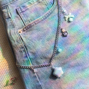 May include: A silver-toned metal chain accessory featuring multiple charms, including a large iridescent star, a small star, a bow, and several round beads, attached to a pair of blue denim jeans with a multi-coloured tie-dye pattern throughout.