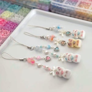 May include: A collection of handmade phone charms with various bead designs. Each charm features a silver-tone wire loop, colorful beads, and a small, white, bear-shaped charm. The beads include pink, blue, and clear colors, with heart and pearl accents.