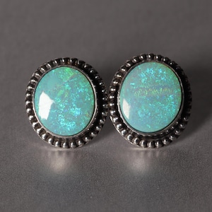 May include: A pair of oval-shaped stud earrings. Each earring features a light blue opal stone with iridescent green and blue flashes, set in a silver-toned, beaded frame. The earrings are displayed on a gray surface.