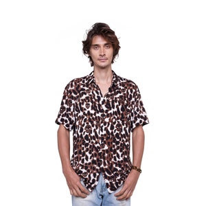 Puede incluir: Camisa de manga corta con estampado de leopardo marrón y blanco y un diseño de calaveras repetido. La camisa está abierta en el cuello. La camisa se lleva con vaqueros azules.