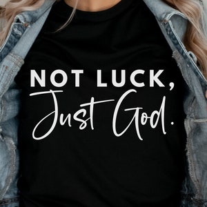May include: Black t-shirt with the white text "NOT LUCK, Just God." The shirt is being worn with a denim jacket. The text is in a bold, sans-serif font.