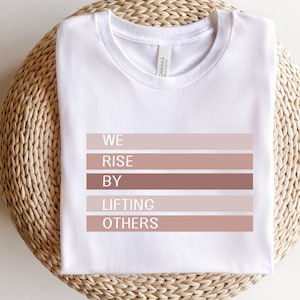 May include: White t-shirt with the text "WE RISE BY LIFTING OTHERS" printed in black on a series of horizontal brown and beige stripes.
