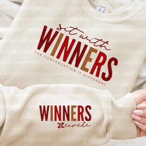 May include: A beige sweatshirt with the text "sit with WINNERS THE CONVERSATION IS DIFFERENT" in red and brown lettering. The bottom of the sweatshirt has the text "WINNERS circle" in red and brown lettering.