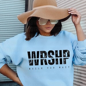 May include: Light blue sweatshirt with the word "WORSHIP" in bold black letters above the words "WHILE YOU WAIT". The sweatshirt is long-sleeved and has a crew neck. The person in the image is wearing a wide-brimmed hat and sunglasses.