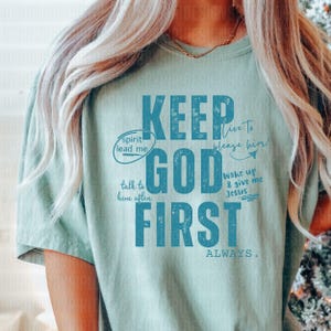 May include: A light green t-shirt with a blue graphic that says "Keep God First Always". The graphic also includes the phrases "spirit lead me", "talk to him often", "live to please him", and "wake up & give me Jesus".