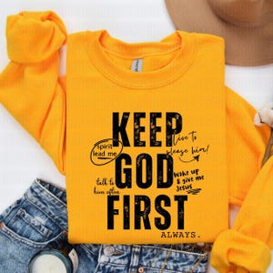 May include: A yellow sweatshirt with the text "KEEP GOD FIRST ALWAYS." in black. The text is in a distressed font and includes additional text in smaller font around the main text, such as "spirit lead me", "talk to him often", "live to please him!" and "wake up & give me Jesus".