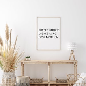 Op de afbeelding: Een witte muur met een ingelijst print waarop staat "Coffee Strong Lashes Long Boss Mode On". Een houten consoletafel met een witte lamp, een plant en manden.