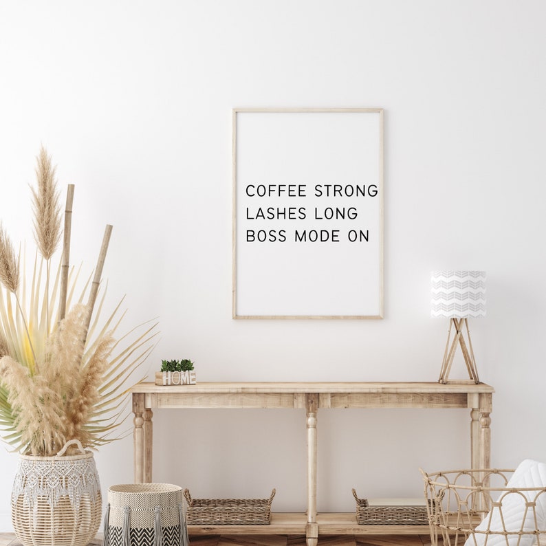 Op de afbeelding: Een witte muur met een ingelijst print waarop staat "Coffee Strong Lashes Long Boss Mode On". Een houten consoletafel met een witte lamp, een plant en manden.