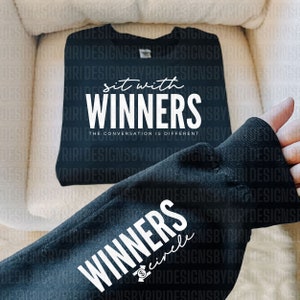 May include: Black t-shirt with white text that reads "Sit with Winners. The conversation is different."  Another black t-shirt with white text that reads "Winners circle" is partially visible.