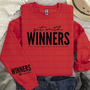 May include: Red sweatshirt with the text "Sit with Winners" and "The Conversation is Different" printed on the front. The left sleeve has the word "Winners" and the word "Circle" printed below it.