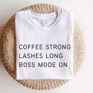 Op de afbeelding: Wit T-shirt met de tekst "COFFEE STRONG LASHES LONG BOSS MODE ON" erop gedrukt.