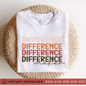May include: White t-shirt with the word "DIFFERENCE" stacked four times in a gradient of colors from light peach to dark brown, with "maker" in a script font. The shirt is folded and placed on a woven, light brown surface.